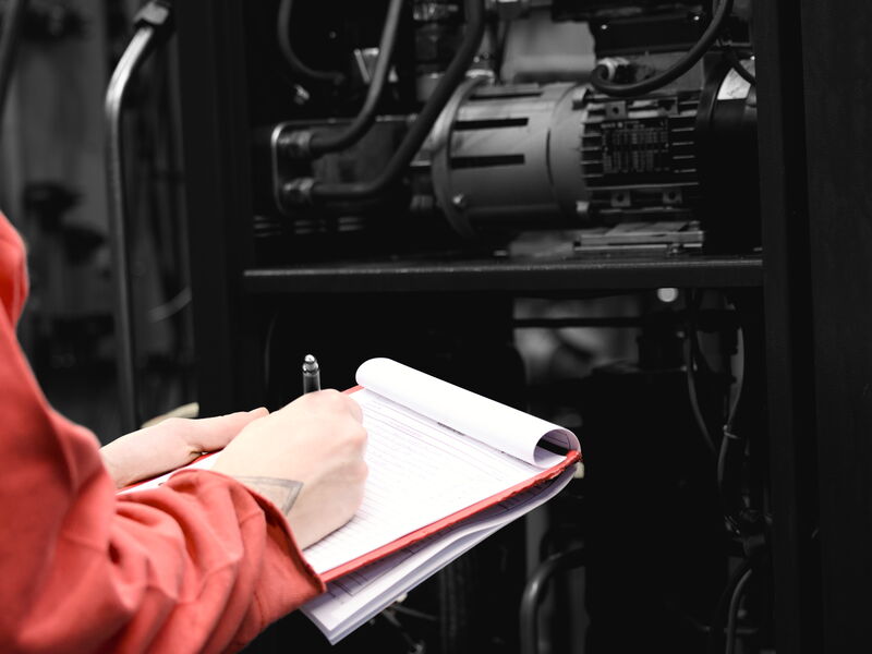 Eine Person in roter Arbeitskleidung schreibt auf einem Klemmbrett, im Hintergrund eine Maschine mit Motor